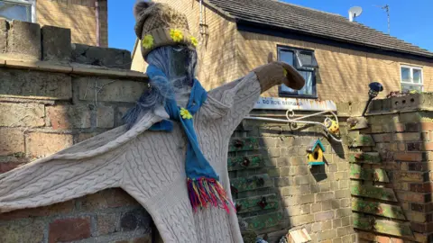 Shariqua Ahmed/BBC A wooden scarecrow that looks like a woman - wearing a beige jumper and a beige woolly hat with yellow flowers.