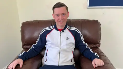 A 29‑year‑old ketamine user discussing bladder damage caused by long‑term recreational ketamine use. Peter is sitting on a brown leather armchair against a cream wall. He is wearing a blue white and red Adidas zip-up jacket. He has light brown hair in a quiff and blue eyes and is smiling at the camera.