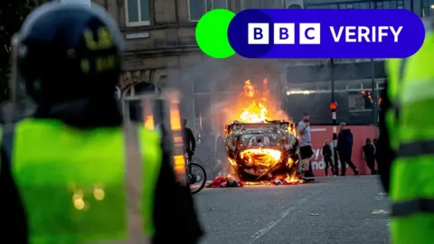 Getty Images Police officers wearing riot gear in Sunderland, viewed from behind close to the camera, as in the middle distance an overturned car burns and a crowd mills around