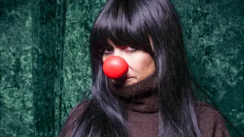 Claudia Winkleman played by Anna Friel, who is wearing a brown fringed jumper, a red nose and a black wig with a blunt fringe.