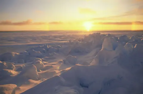 Getty Images - Doug Allan Sun setting over a frozen sea, illuminating jagged ice formations and snow-covered pressure ridges beneath a golden sky.
