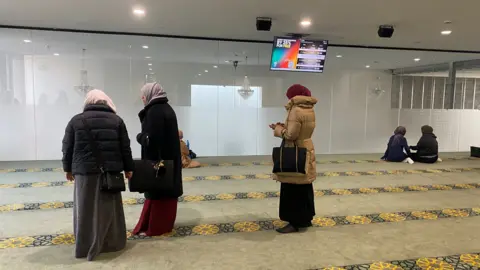 A large room with a carpet has a number of Muslim women sitting and praying together. A group of three are standing in the forefront of the picture 
