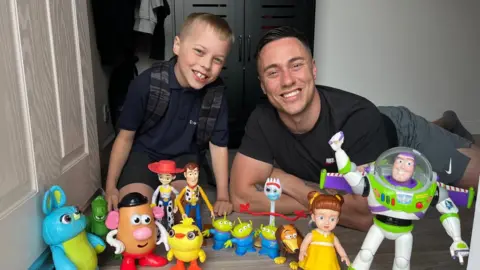A child and his father lying at a doorstep with an array of toys from the Toy Story film franchise
