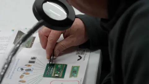 Jamie Niblock/BBC A person is using a pair of tweezers to move components on a small green board. There are instructions on a page and the person is using a magnifying glass to help them see the tiny parts.