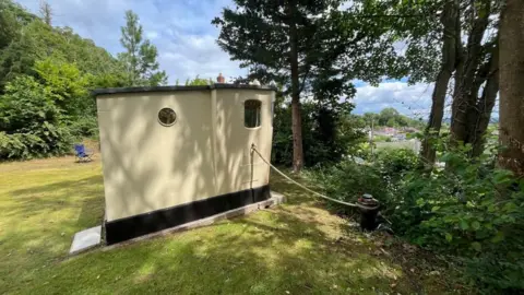 Ellen Knight/BBC The shed, as restored by Richard and Abi. It has cream walls and a black roof, with small circular windows on the sides and larger rectangular windows on its from. There is a rope, tied on to a mooring post, and it's surrounded by grass and shrubbery. It looks out over a small ridge onto hills and more countryside. The sky is blue with some clouds. 