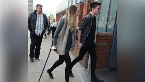 PA Media A girl wearing a furry gilet with blond hair and a grey jumper is aided by a walking stick. She is holding the had of a young man in a checked shirt. A second man wearing a dark jacket and light shirt walks behind them