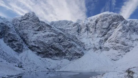 SAIS Creag Meagaidh The craggy mountainside is dusted with snow. There is a lochan below the crags and wispy white cloud above the summits.