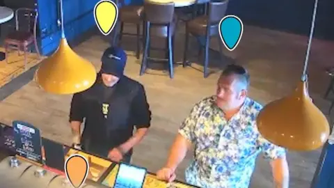 Devon and Cornwall Police Still from CCTV footage showing two men stood at the bar of a pub. The man on the left is wearing dark clothing and a baseball cap. The man on the right has a white and blue floral shirt on. He has a moustache.