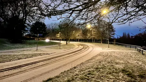 SignmakersWife/WEATHERWATCHERS A snow-covered road curves around a corner. It is dusk. A row of streetlights are lit. Grass verges on wither side are covered in frost and snow.