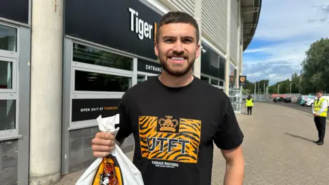 Matt Dean/BBC Jay King pictures with a Hull City carrier bag outside the stadium. He is wearing a black t-shirt and had short dark hair and a beard.