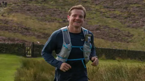 A man running in green scenery. He is smiling and looking at the camera.