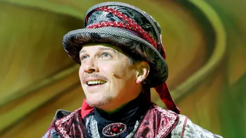 A head and shoulders image of Nigel Harman shows him in a black and red Fleshcreep costume. He wears a black top hat style hat with red braiding and his shiny black coat has a red collar. He has short, dark brown hair with a small moustache and beard and green eyes. He has a head microphone resting on his cheek and is speaking out to the audience.