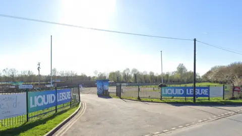 A Google Streetview of a road heading to a car park with a Liquid Leisure sign on a blue hoarding attached to the fence alongside.