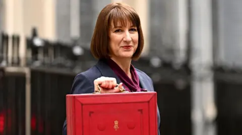 Chancellor Rachel Reeves is looking into the camera outside Downing Street. She is wearing a purple top, a blue blazer and is holding the red Budget box.