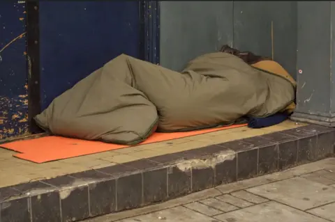 A figure in a brown sleeping back on a tiled step