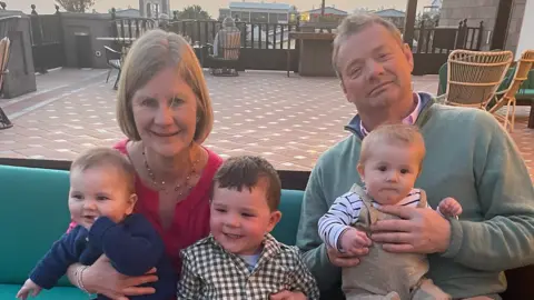 A woman with blonde hair and a red dress, along with a fair-haired man in a green jumper sit with three toddlers on their knees.