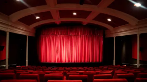 Kat Gollock A view of the red-curtained big screen from the back of screen one, the rows of red seats stretching down to the screen.