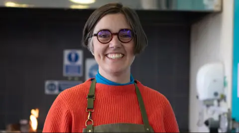 Pictured is Rebecca Skeels smiling in a classroom. She has cheek length dark and grey hair and is wearing thick round glasses. She has on a red high neck jumper with a blue roll neck sweater underneath and a khaki apron.
