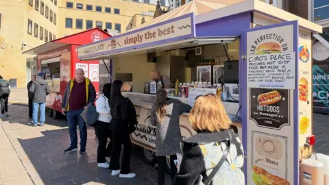 Ollie Conopo/BBC Several people queue outside of a food van. The van is branded CIRCO'S PLACE - SIMPLY THE BEST. It is a sunny day.
