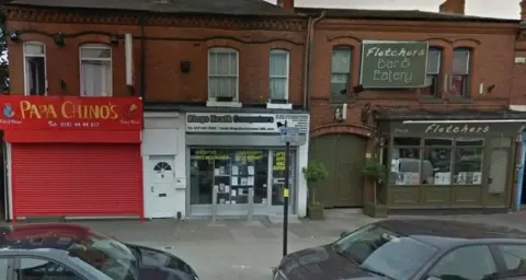 Google A series of terraced shops and flats can be seen from across the road. One red shuttered shop is called Papa Chino's, and two doors down is Fletchers Bar & Eatery. A couple of cars can be seen parked next to the pavement.