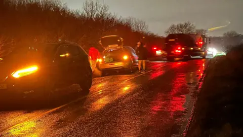 Becky Broadley A row of cars on a road, it is dark so you can only make out some headlights, some blurred images of cars and a few people standing by them. 