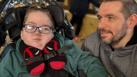 Stephen Browne has blue eyes and short grey hair. He is wearing a grey hoodie. His son Milo is next him. His blonde hair is put up in a small bun. He has blue-rimmed glasses and is wearing a green hoodie.