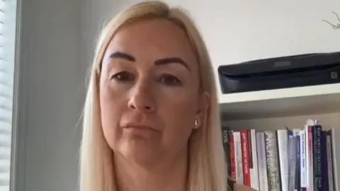 A woman with shoulder-length blonde hair is in front of a book shelf with several books on it in front of a white wall and a blind.