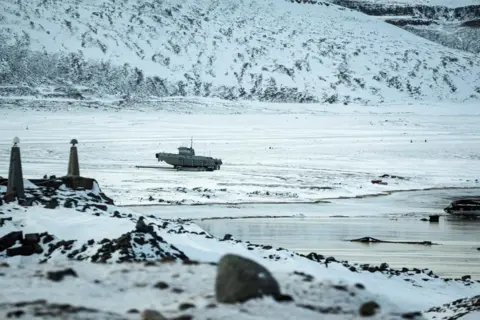 Frozen water and a snow-covered landscape in Greenland, with a military-looking vehicle pictured in the middle distance