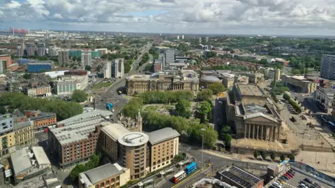 An aerial view of Liverpool city centre looking toward St George's Hall, St John's Gardens and William Brown Street. The River Mersey is in the distance.