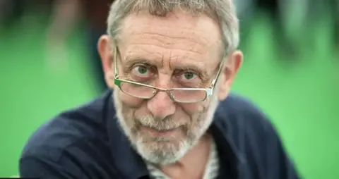 Getty Images Michael Rosen is wearing glasses and gazing into the camera. His face is in focus, but his blue jacket and a green background behind him are blurred.