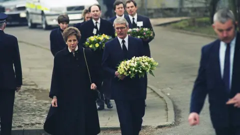 Getty Images A group of seven people walk towards camera down a street with police cars in the background. John Major, Michael Forsyth and Tony Blair can be seen carrying wreathes. George Robertson can be seen in the background. All are in dark suits, white shirts and black ties. 