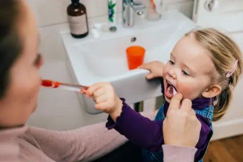 A little girl is standing by a sink with a woman standing next to her. They are brushing each other's teeth. 