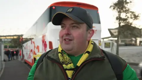 Ian Lobb stands in front of a white coach outside Plymouth Argyle's Home Park stadium. He is wearing a grey baseball cap, a yellow and green scarf, a green Argyle football shirt and a green gilet.