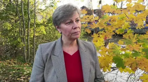 A woman in a red top and grey blazer standing outside next to a river. She has short grey hair and is looking to the left.