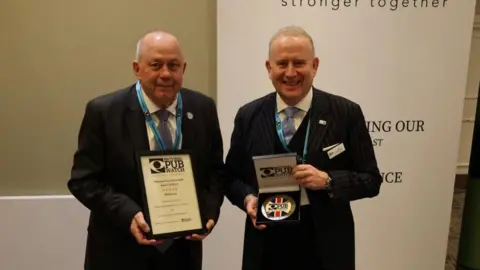 Two grey-haired men wearing suits and smiling at the camera. The one on the left is holding up a certificate and the one on the right is holding a plaque. Both items have the National Pubwatch logo on them