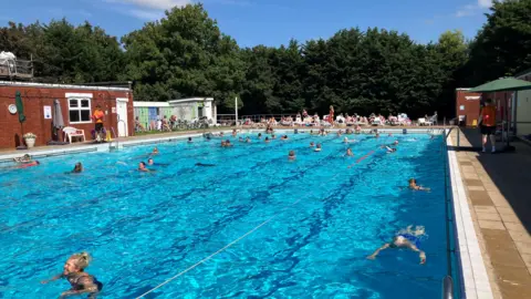 A large blue-coloured outdoor swimming pool, with light glistening on the water. There are people in the water, lifeguards on the sides of the pool, and other people sat on sun loungers