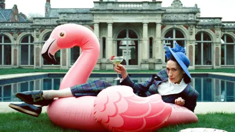 Metfilm Sian Clifford lounges on the grass in front of a large square pond with part of Somerleyton Hall behind her. She wears a checked suit, a blue hat and is holding a cocktail glass in one hand, while propping herself up on one elbow, and leaning against an inflatable pelican in front of her on the lawn. 