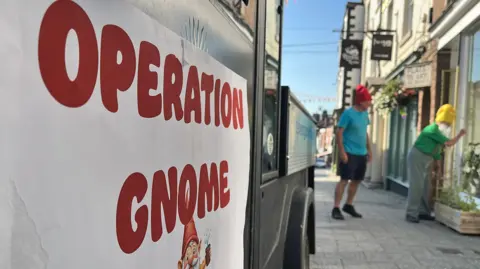 Ellen Knight/BBC A close-up photo of the 'Operation Gnome' sign - red text on a white background with a small cartoon gnome underneath. The sign is in focus - with two volunteers out of focus beyond it. The sky is bright blue and the high street has lots of sunshine. 