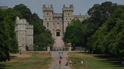 PA View of a large stone castle at the end of a broad, tree-lined avenue. A paved path runs through a grassy park toward the castle’s arched entrance, with several visitors walking, crossing the lawn, and pushing a stroller in the foreground. The castle features tall square towers, battlements, and Gothic-style windows, with a smaller stone tower visible to the left. Mature trees frame both sides of the scene, and the building rises above the surrounding greenery under a clear sky.