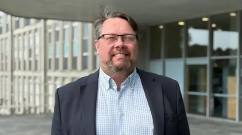 Dan has brown hair and a beard with glasses and wears a white and blue checked shirt and dark jacket. He stands in front of the entrance to Lys Kernow. 