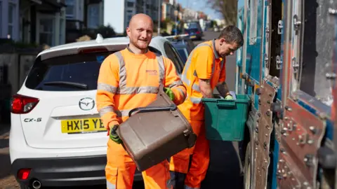 Bristol Waste Company Two male waste collectors dressed in high-vis orange uniform. One is holding a brown food caddy and is smiling to camera. The other has a green recycling box balanced up against a refuse truck. There is a white car behind them