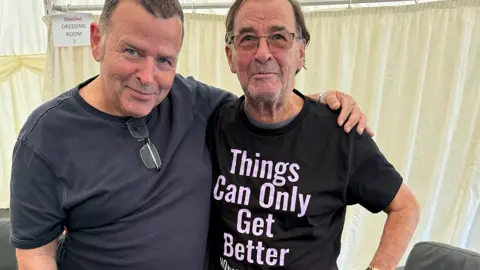 Peter Cunnah On the left is a man in his 50s wearing a grey t-shirt with glasses hanging from the neck of the garment. He has his left arm round an older man who is wearing a black t-shirt with the logo "Things Can Only Get Better".
