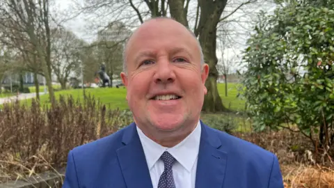 Jim O'Boyle, cabinet member for jobs at Coventry City Concil smiles at the camera. He is wearing a blue suit jacket, white shirt and blue tie. Behind him we see trees, grass and bushes.