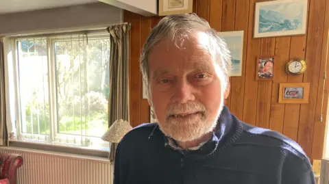 James Grant/BBC A man in a navy zip-up jumper standing in a living room with a wooden wall behind him. He is looking at the camera.