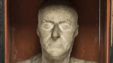 James Sinclair/Fitzwilliam Museum Clay mask of a face, with eyes shut and mouth closed, photographed in front of a red background.