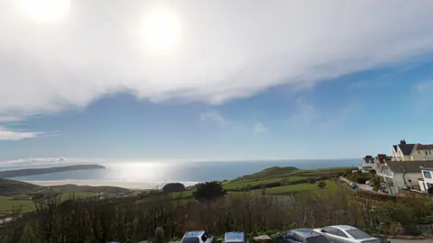 A scenic coastal photo looking out from a hotel balcony with the sea in the distance. Bright blue skies with the blue sea below. There is lots of green fields as well as some buildings to the right hand side of the photo. There are also a few cars parked and a sandy beach in the background.