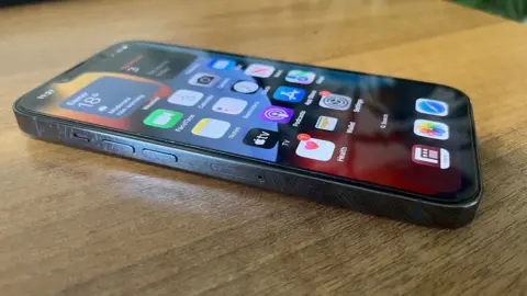 A black iPhone on a desk with various apps on the home screen.