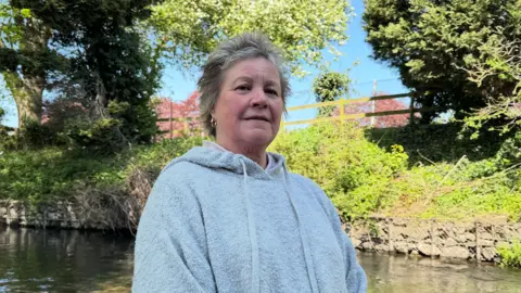 Tessa Smith-Winnard is wearing a grey hooded top and standing in front of a brook.  She has short, grey hair. 