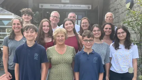 Joan Simon is in the front row of a family photograph with her two grandchildren dressed in blue polo tops with the school logo in white on them. They are flanked by other family members who are all standing outside the entrance to the school, with 14 people in total. A sign reading Welcome to Les Landes School can be seen above the front door.