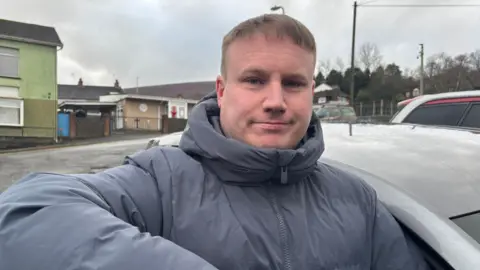 A man is seen leaning on his car door. He looking at the camera and wearing a navy blue coat which is zipped to the top. He has sand colour hair. He's standing in a small car park and behind him there's a social club.